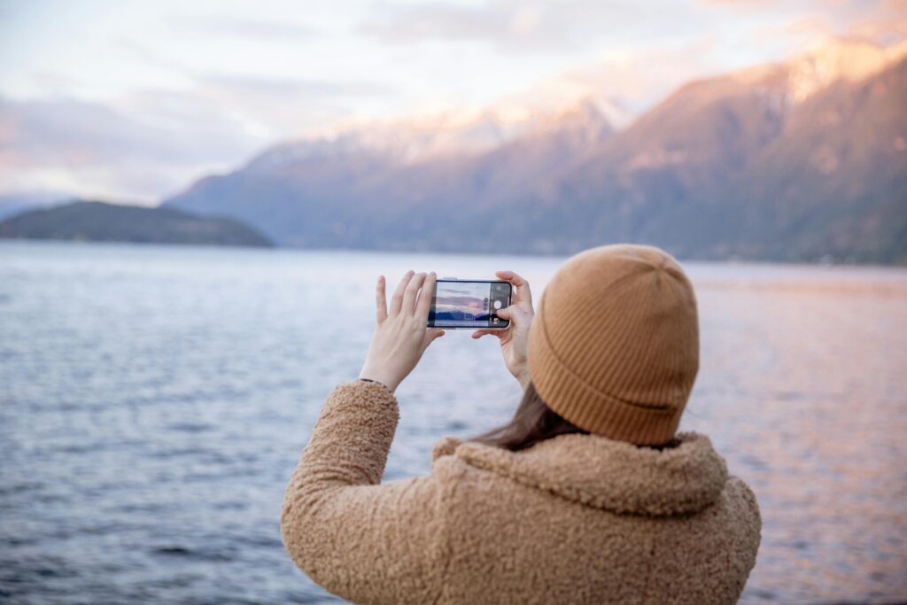Back view of unrecognizable female tourist in warm coat and hat taking photo on smartphone while standing on coast near water and enjoying seascape during trip