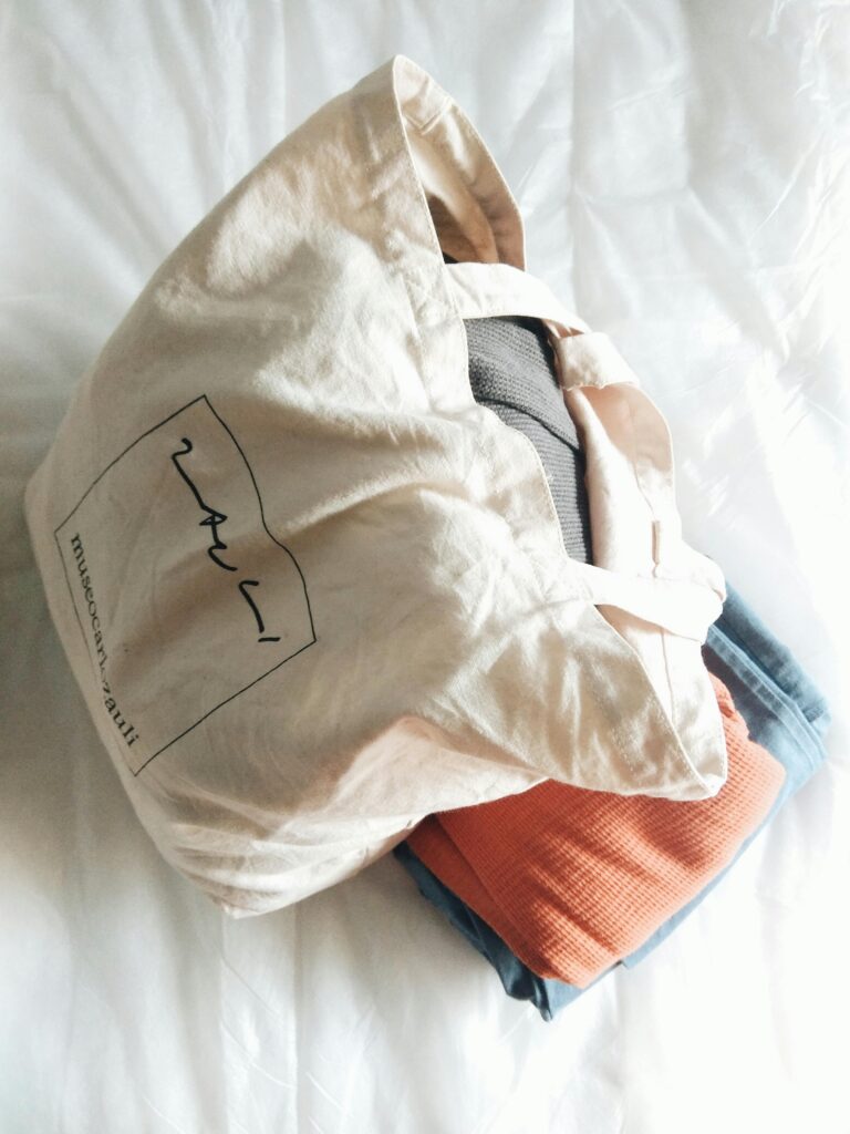 A natural cotton tote bag filled with clothes resting on a neatly made bed indoors.
